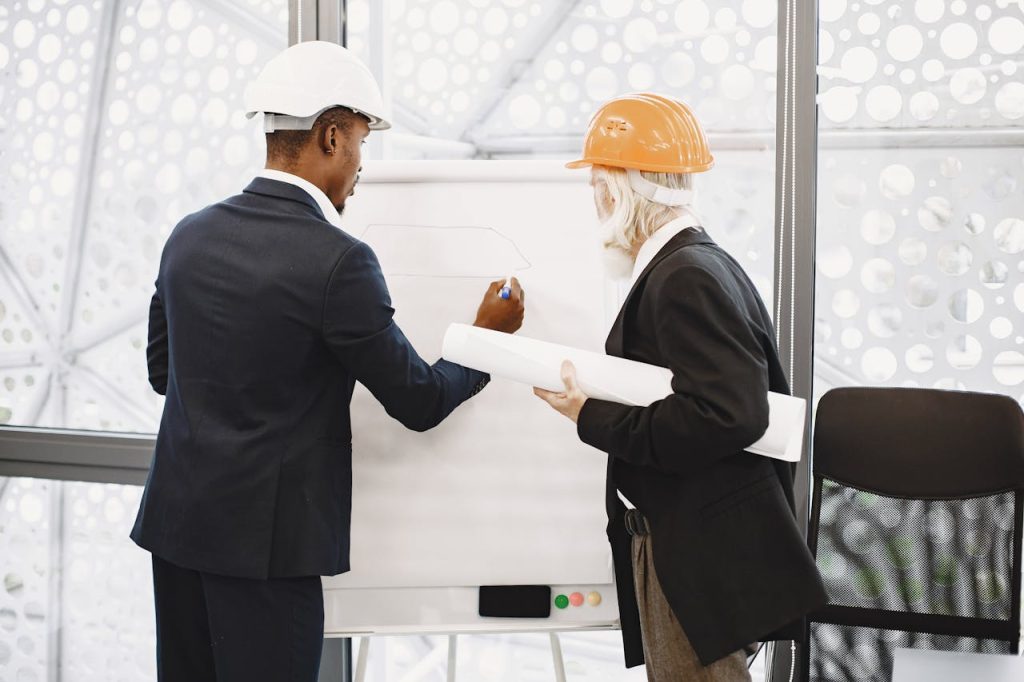 Two architects in hard hats working on a blueprint indoors, highlighting teamwork.
